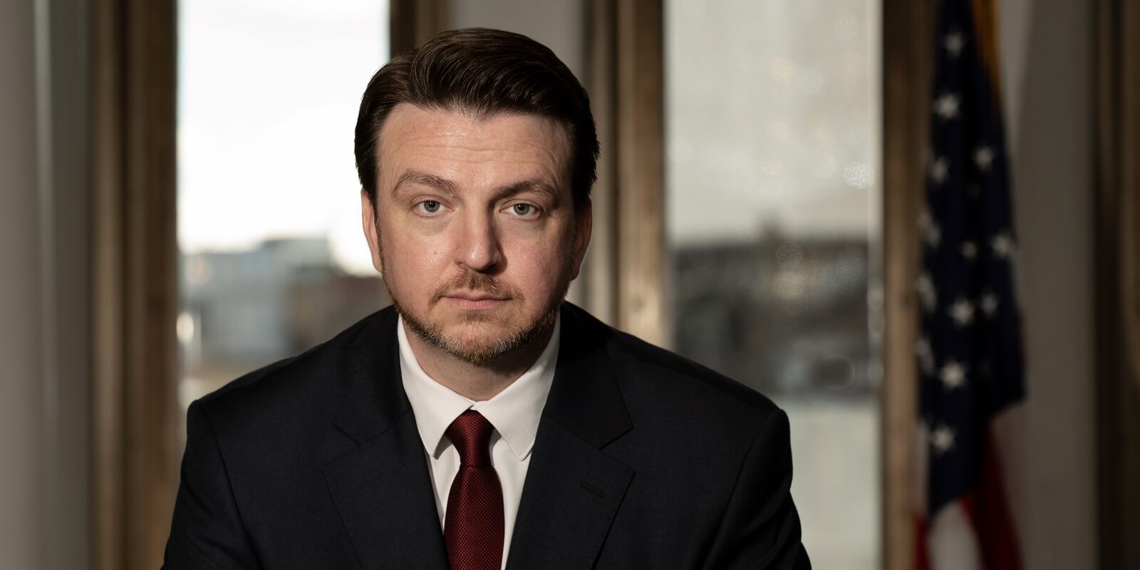 Philip Foust in a dark suit and red tie beside an American flag, a downtown Indianapolis window behind him.